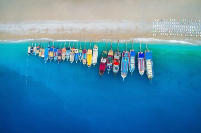 Aerial view of colorful boats tied side by side along a sandy beach, with bright blue water fading to deep blue.
